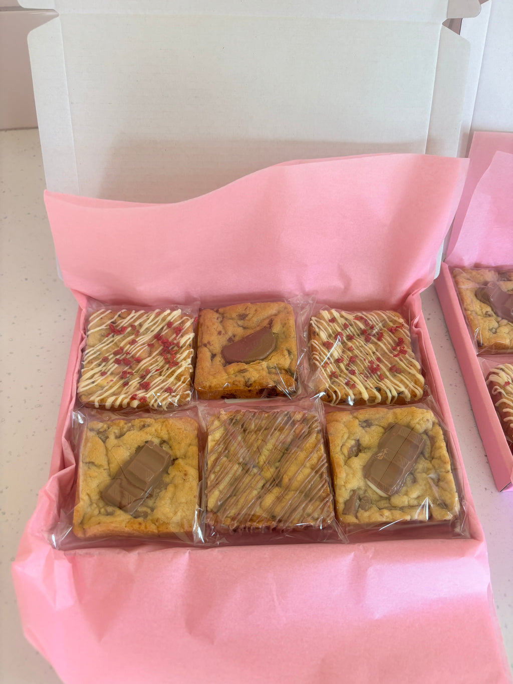 An open pink gift box containing six individually wrapped square cookies arranged in two rows, with pink tissue paper lining the box.